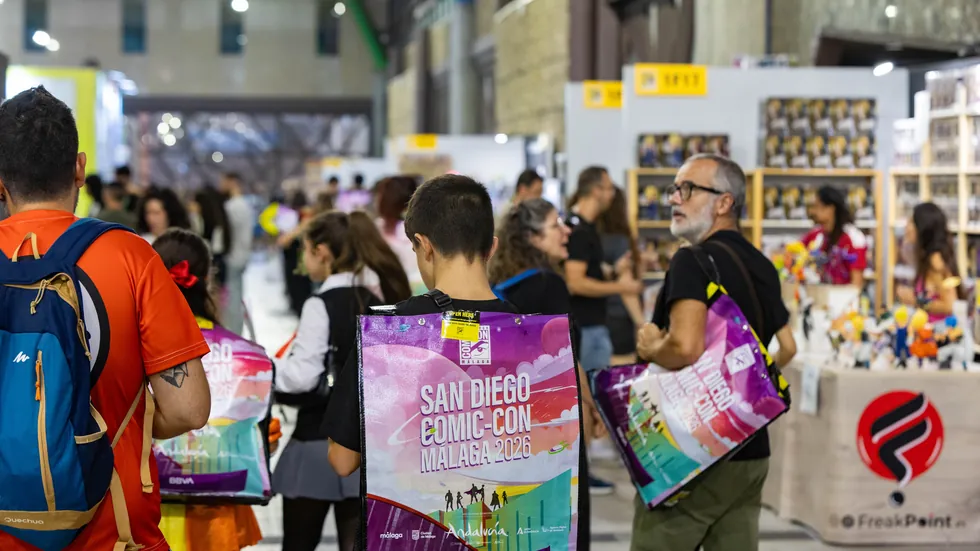 Pasillo interior del recinto con stands y asistentes cargando la bolsa oficial del evento